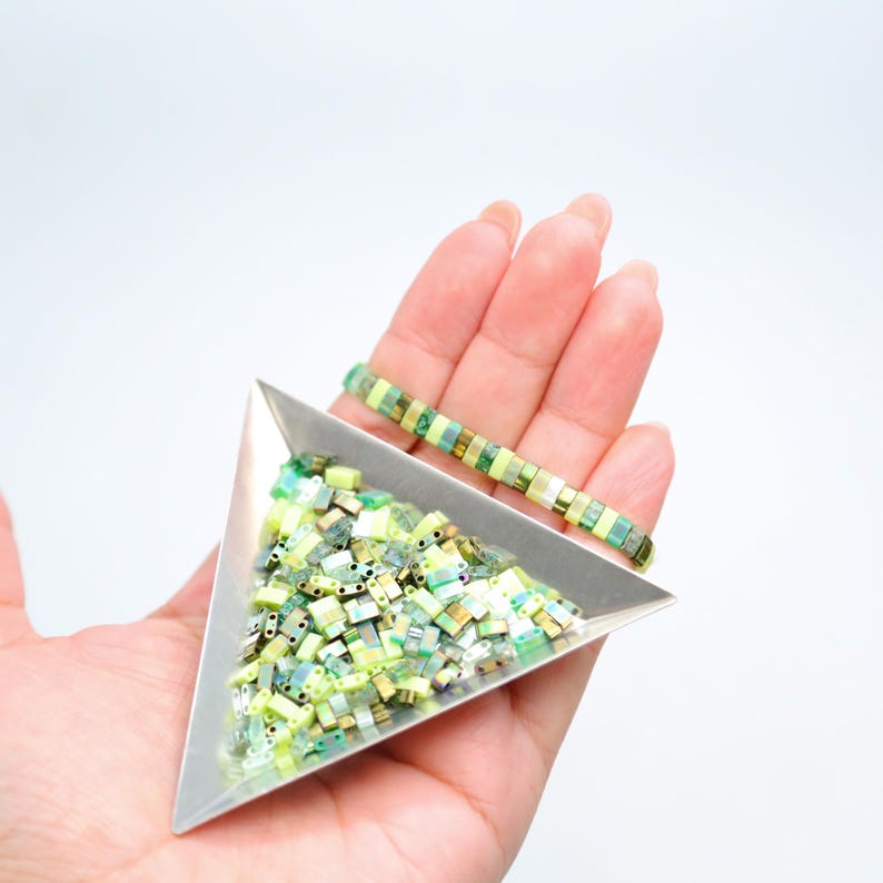 May include: A triangular tray filled with green and yellow square beads. A bracelet made of the same beads is lying on top of the tray.