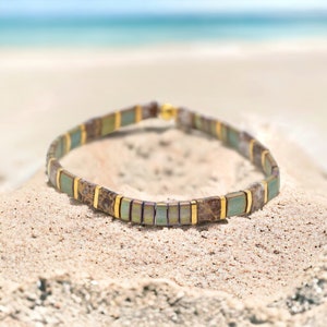 May include: A brown and green beaded bracelet with gold accents. The bracelet is made of small, square beads and is lying on a sandy surface.