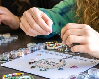 May include: A person is making a bracelet with colorful beads on a white mat with the words "& REX MAGIC" printed on it. There are small containers of beads in various colors around the mat.