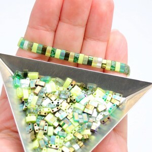 May include: A close-up of a tray of green and gold iridescent glass beads. The beads are in the shape of small rectangles. A bracelet made with the same beads is shown in the background.