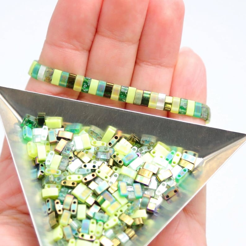 May include: A close-up of a tray of green and gold iridescent glass beads. The beads are in the shape of small rectangles. A bracelet made with the same beads is shown in the background.
