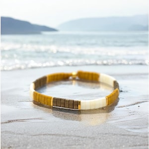 May include: A colorful beaded bracelet with gold accents. The bracelet is made of brown, white, and yellow beads. The bracelet is lying on a sandy beach.
