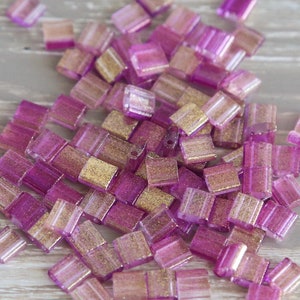 May include: A collection of small, square beads in shades of pink and gold. The beads have a translucent quality, with a glittery gold interior. These beads are ideal for jewelry making and other craft projects.
