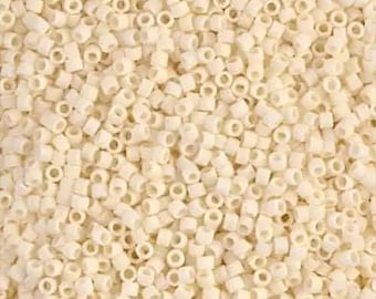 Op de afbeelding: Een close-up van een stapel kleine, crèmekleurige, cilindrische kralen. Elke kraal heeft een gat in het midden. De kralen zijn verspreid, waardoor een textuur ontstaat. Deze kralen zijn geschikt voor het maken van sieraden en knutselen.