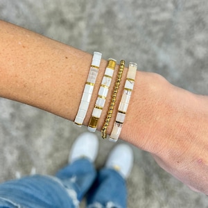 May include: Four bracelets stacked on a wrist. The bracelets are made of white beads with gold accents. The bracelets are a mix of round and square beads.