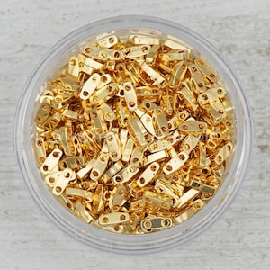 May include: A close-up view of a container filled with gold-colored metal jump rings. The jump rings are small and oval-shaped with a hole in each end.