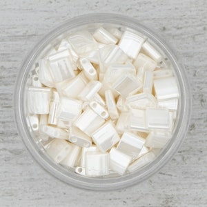 May include: A close-up view of a clear glass jar filled with white iridescent square beads. The beads have a smooth, glossy finish and are arranged in a random pattern.