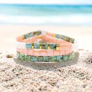 May include: A set of three bracelets with square beads in shades of pink, white, and green. The beads have a mottled, textured appearance. The bracelets are stacked on top of each other, creating a layered effect.