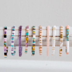May include: A collection of colorful beaded bracelets with various patterns and colors, including pink, purple, turquoise, white, and gold. The bracelets are displayed on a white wooden surface.