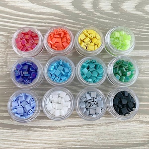 May include: Twelve small containers filled with colorful square beads. The containers are arranged in three rows of four. The beads are in the colors red, orange, yellow, green, blue, teal, white, gray, and black.