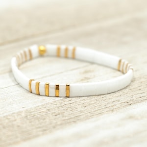 May include: White bracelet with gold accents. The bracelet is made of small, white ceramic tiles with gold accents.