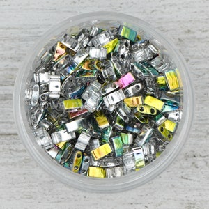 May include: A clear plastic container filled with small, rectangular beads. The beads are silver with iridescent, rainbow-colored surfaces. The beads have two holes for stringing. The container sits on a light-colored, textured surface.