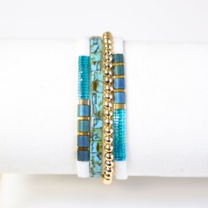 May include: A set of five bracelets featuring a mix of turquoise and gold beads. The bracelets are stacked on a white surface.