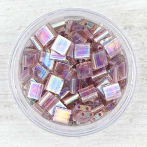 May include: A close-up view of a clear container filled with iridescent pink and purple glass beads. The beads are small and rectangular in shape, with a slightly rounded edge. The beads have a shimmering, iridescent finish that reflects light in a variety of colors.