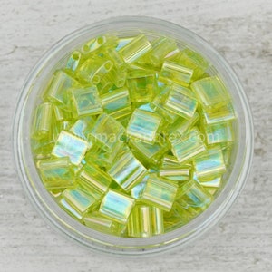 May include: A glass jar filled with small, iridescent green glass beads. The beads are square shaped with a slightly rounded edge.