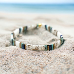 May include: A white and blue bracelet with gold accents. The bracelet is made of small, square tiles and is lying on a sandy beach.