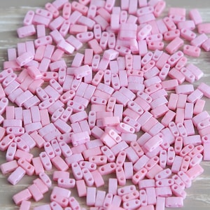 May include: A collection of small, rectangular, light pink beads with two holes each. The beads are scattered across a light-colored surface, creating a textured appearance. These beads are likely used for jewelry making or other craft projects.