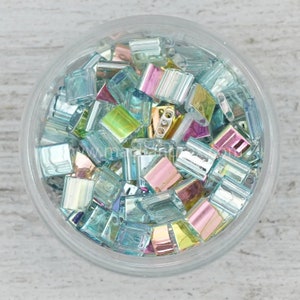 May include: A clear plastic container filled with iridescent, rectangular beads. The beads have a mix of colors, including blue, pink, and yellow. The beads are small and appear to be made of glass or a similar material. The container is sitting on a white surface.
