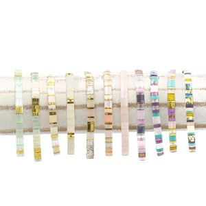 May include: A collection of 12 colorful beaded bracelets with gold accents. The bracelets are arranged in a row on a white striped surface.