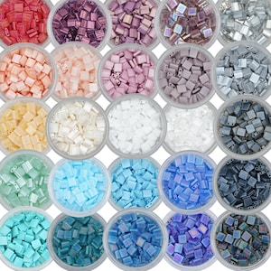 May include: Assortment of 24 small square glass beads in various colors and finishes, including iridescent, opaque, and transparent. The beads are arranged in a grid pattern in small containers.