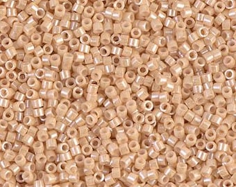 Op de afbeelding: Close-up van een stapel kleine, cilindervormige kralen in een lichtbeige kleur. De kralen hebben een subtiele, iriserende glans die het licht reflecteert. De kralen zijn uniform van grootte en vorm, met een gat in het midden. Ideaal voor het maken van sieraden en knutselen.