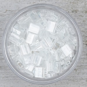 May include: A clear plastic container filled with small, clear, square-shaped beads. The beads are translucent and have a slight shimmer.
