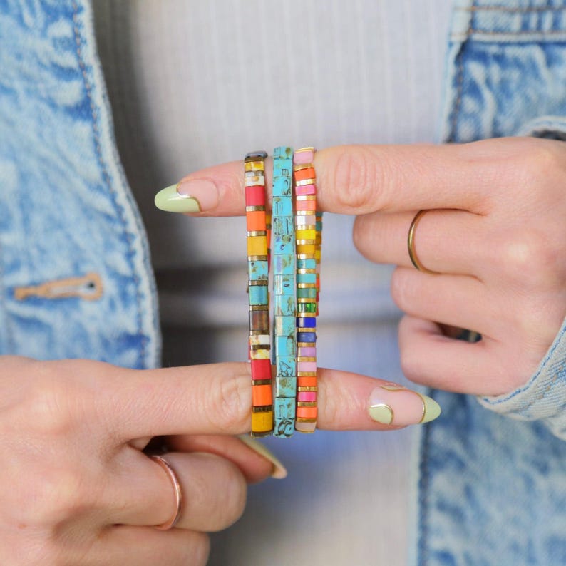 May include: Four colorful tile bracelets stacked on a finger. The bracelets are made of small, square tiles in various colors, including turquoise, pink, yellow, orange, and blue.