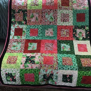 May include: A colorful patchwork quilt featuring a watermelon theme. The quilt is composed of various squares in shades of green, pink, and red, with watermelon slice patterns. It has a black border with a pink trim.