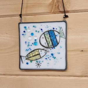 May include: Square glass art piece with two stylized fish in black outlines, with blue and yellow accents. The background is white with blue and clear glass pieces, and a black cord for hanging.