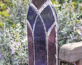 May include: A stained glass window hanging ornament in a gothic arch shape. The glass is a deep purple and burgundy, with silver glitter accents. The ornament hangs from a silver chain, with a natural background of green foliage.