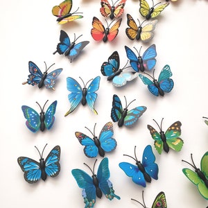 May include: A collection of colorful butterfly decorations in various sizes and designs. The butterflies feature shades of blue, green, yellow, orange, and pink, with black antennae and bodies. They are arranged on a white surface.