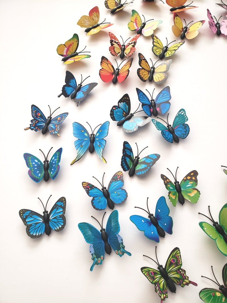 May include: A collection of colorful butterfly decorations in various sizes and designs. The butterflies feature shades of blue, green, yellow, orange, and pink, with black antennae and bodies. They are arranged on a white surface.