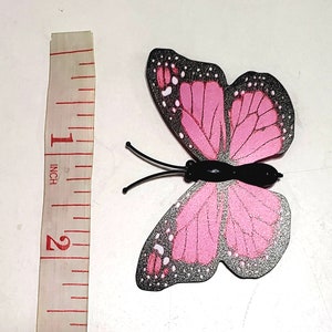 May include: A decorative butterfly with pink wings and black accents. The butterfly is approximately 2 inches in length, with a black body and antennae. The wings have a detailed pattern with white dots.