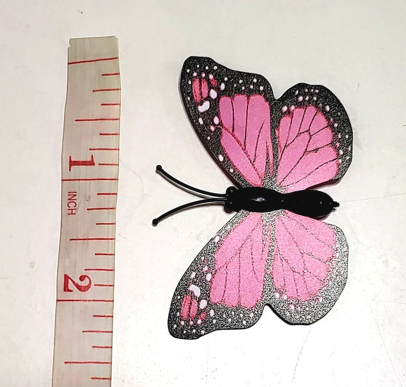 May include: A decorative butterfly with pink wings and black accents. The butterfly is approximately 2 inches in length, with a black body and antennae. The wings have a detailed pattern with white dots.