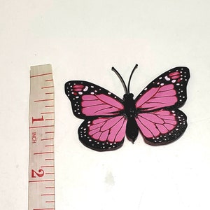 May include: A pink and black butterfly decoration with white accents. The butterfly has a wingspan of approximately 2 inches. The butterfly has black antennae and a black body. The butterfly is on a white background.