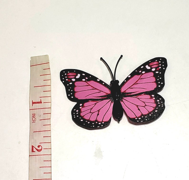 May include: A pink and black butterfly decoration with white accents. The butterfly has a wingspan of approximately 2 inches. The butterfly has black antennae and a black body. The butterfly is on a white background.