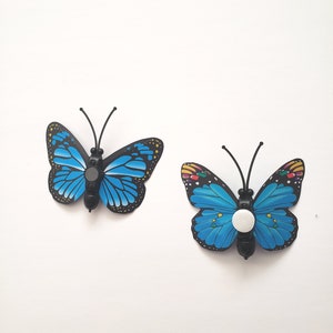 May include: Two decorative butterfly-shaped objects. The butterflies have bright blue wings with black and yellow detailing. Each butterfly has a black body and antennae. One has a white button-like object on its body.