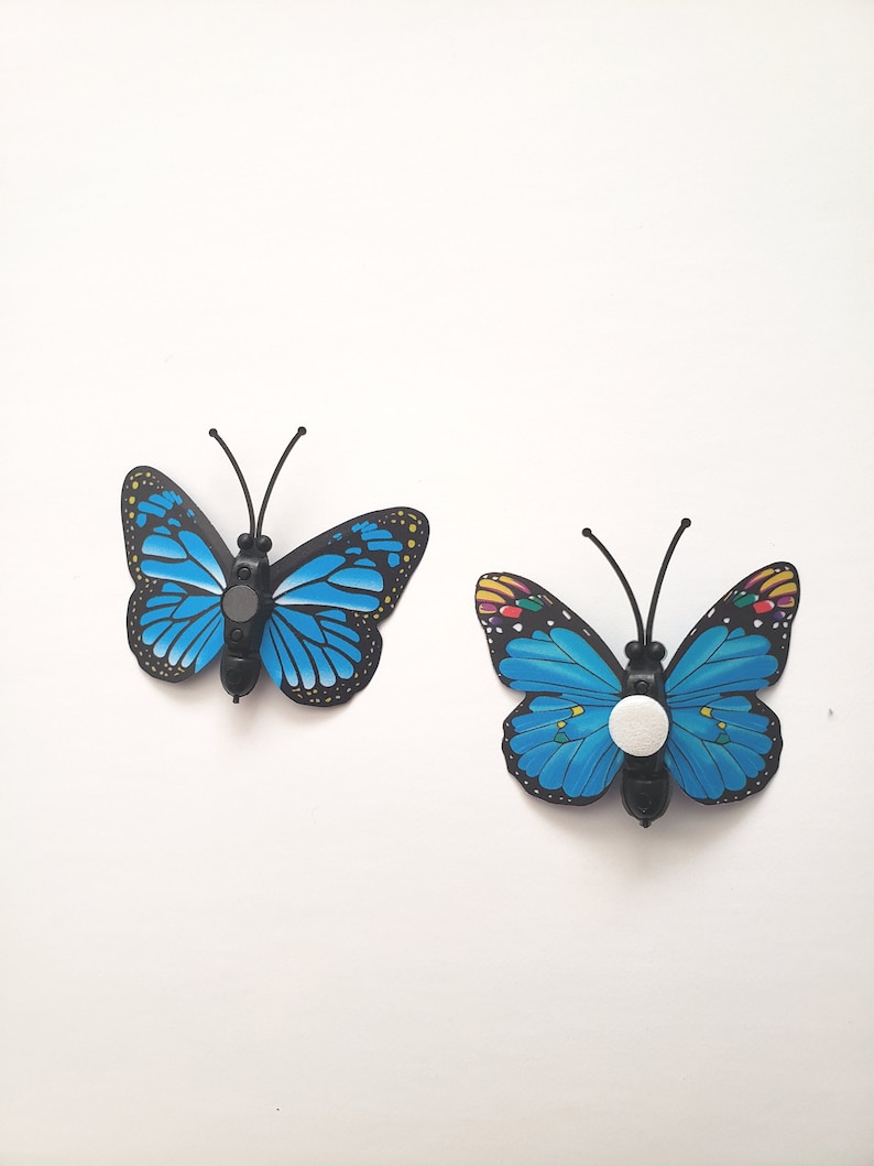 May include: Two decorative butterfly-shaped objects. The butterflies have bright blue wings with black and yellow detailing. Each butterfly has a black body and antennae. One has a white button-like object on its body.
