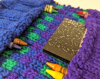 May include: A handmade, knitted pencil case in shades of blue, green, and purple, with a black and white composition notebook and several crayons. The case is designed to hold school supplies.