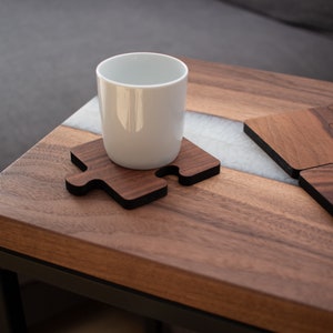 Könnte beinhalten: Eine weiße Keramiktasse steht auf einem Untersetzer aus Holz in Puzzleform. Der Untersetzer befindet sich auf einem Holztisch mit einer weißen Einlage. Der Tisch hat einen schwarzen Metallrahmen.