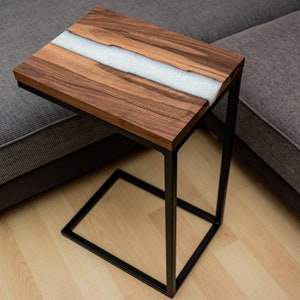 Könnte beinhalten: Ein kleiner Holz-Beistelltisch mit einer weißen Harzeinlage. Der Tisch hat ein schwarzes Metallgestell und steht auf einem Holzboden.