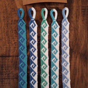 May include: Five friendship bracelets in a chevron pattern with blue, green, and white colours. The bracelets are laid out in a row on a wooden surface.