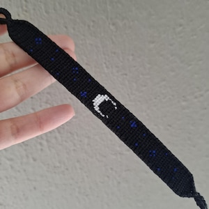 May include: Black woven friendship bracelet featuring a white crescent moon design and small blue accents. The bracelet is handmade and has a knotted closure. The bracelet is a dark color and has a celestial theme.
