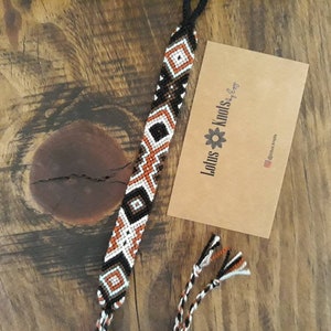May include: A black, white, orange, and brown friendship bracelet with a geometric pattern. The bracelet is on a wooden surface with a business card that says "Lotus Knots by Liz".