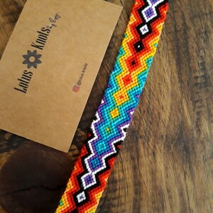 Könnte beinhalten: Ein farbenfrohes Freundschaftsarmband mit einem geometrischen Muster in Rot-, Orange-, Gelb-, Grün-, Blau-, Lila- und Schwarztönen. Das Armband ist mit einer Knotentechnik hergestellt und hat eine schwarze Schnur am Ende.