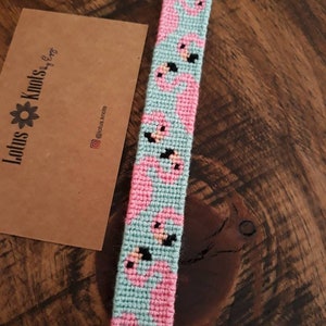 May include: A turquoise and pink friendship bracelet with a pattern of pink flamingos. The bracelet is made with embroidery floss and has a knotted fringe at the end.