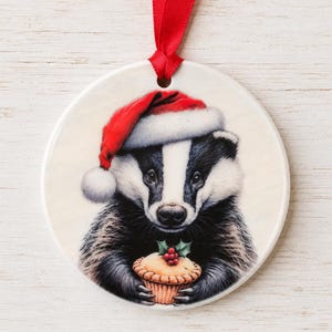 May include: A round white ceramic ornament featuring a black and white badger wearing a red and white Santa hat. The badger is holding a small pastry topped with green holly leaves and red berries. A red ribbon is attached to the top for hanging.