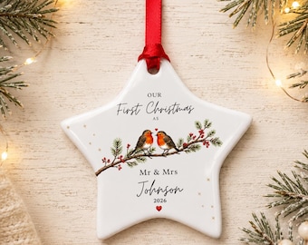 May include: A white star-shaped ceramic ornament hanging from a red ribbon. The ornament features a printed illustration of two robins on a pine branch with red berries, and the text: Our First Christmas as Mr & Mrs Johnson 2026.