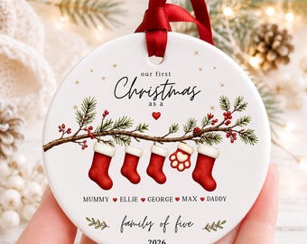 May include: A round white ceramic Christmas ornament with a red ribbon hanger. It features a pine branch with red berries, four red stockings, and one red paw print stocking. Text reads: our first Christmas as a family of five, 2026, and names.