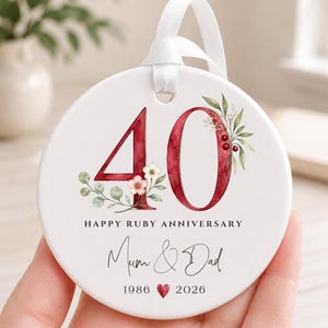 May include: A round white ceramic ornament featuring the number 40 in dark red watercolor, decorated with green leaves and small flowers. It includes the text HAPPY RUBY ANNIVERSARY, Mum & Dad, and the years 1986 and 2026 with a small red heart.